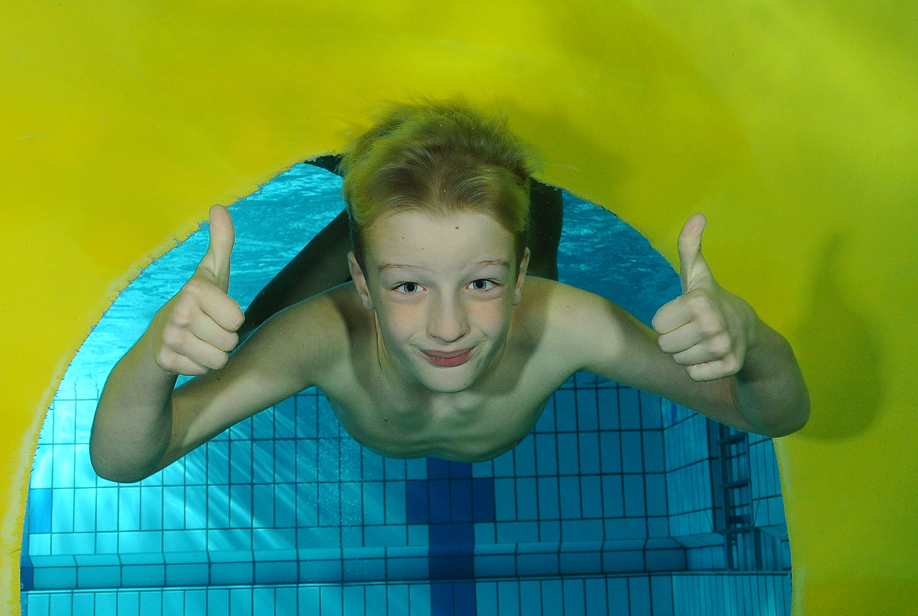 Kind steekt onder water twee duimen op onder de gele glijbaan